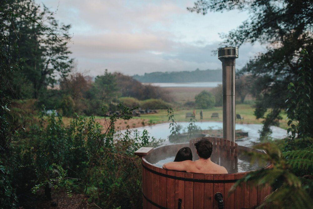 Wooden hot tub with steam vent surrounded by greenery and lake view