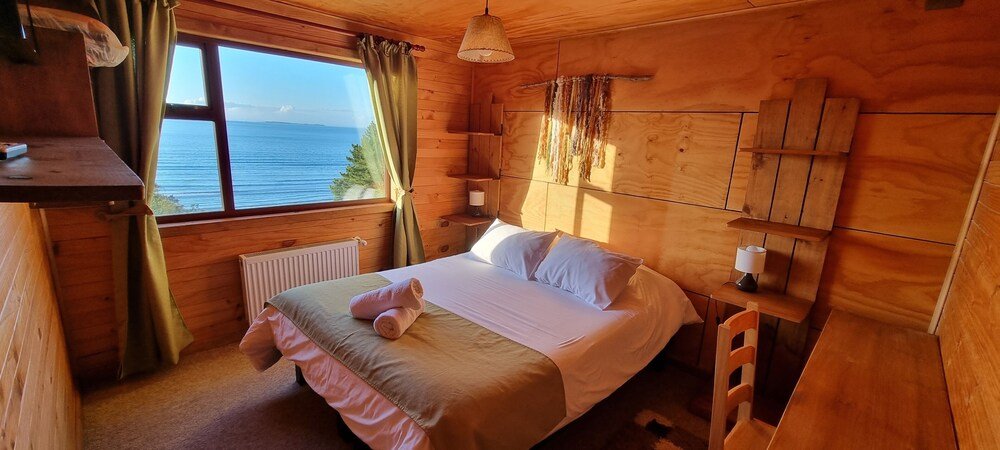 Dormitorio de cabaña de madera con cama, mesa de luz y ventana al mar
