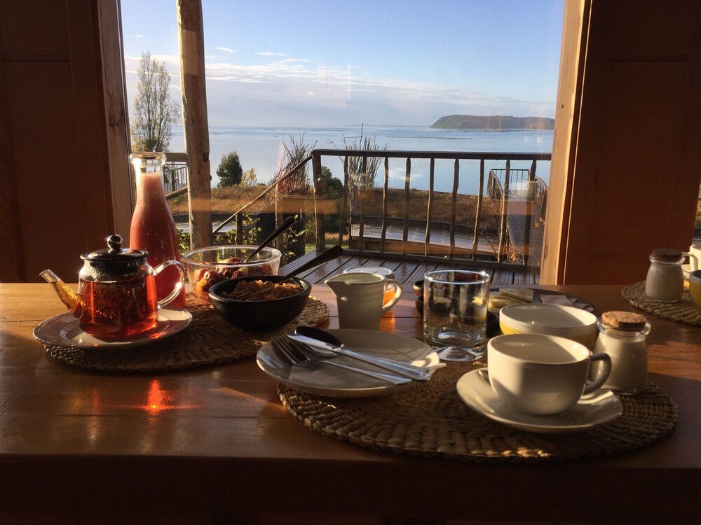 Mesa de desayuno con té, cereales y tazas frente al mar y las colinas